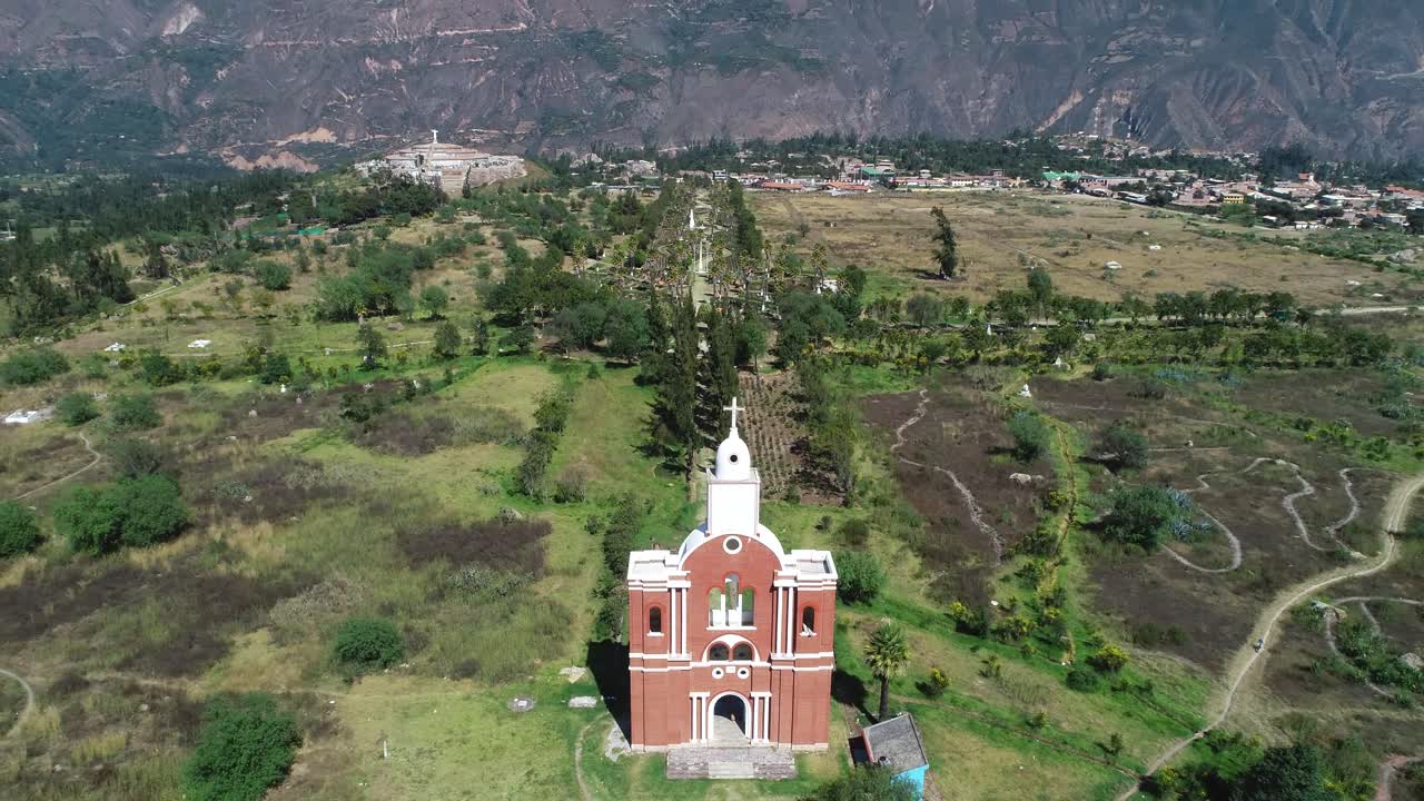 Expansive aerial shot of the church at Yungay Cemetery, Áncash, Peru, surrounded by a vast green valley and Andean mountains. A historic memorial site for the 1970 earthquake victims