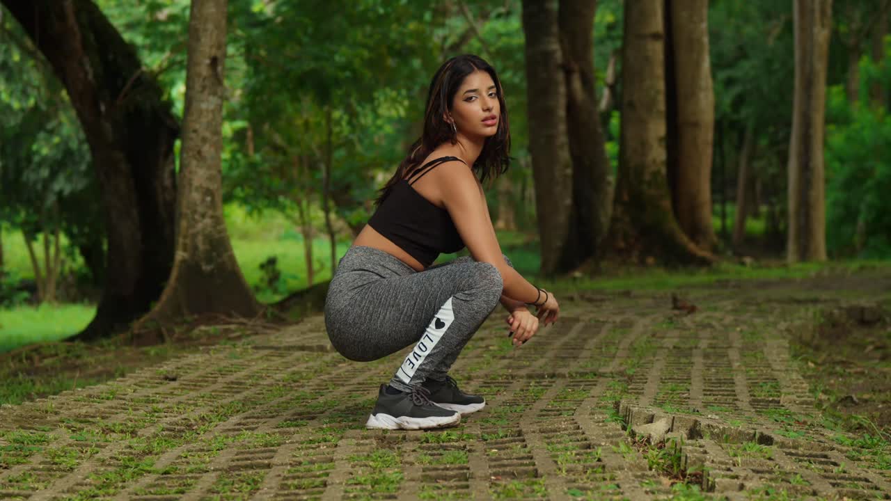 En medio de un parque tropical, una joven se ejercita, vestida con ropa deportiva, haciendo sentadillas