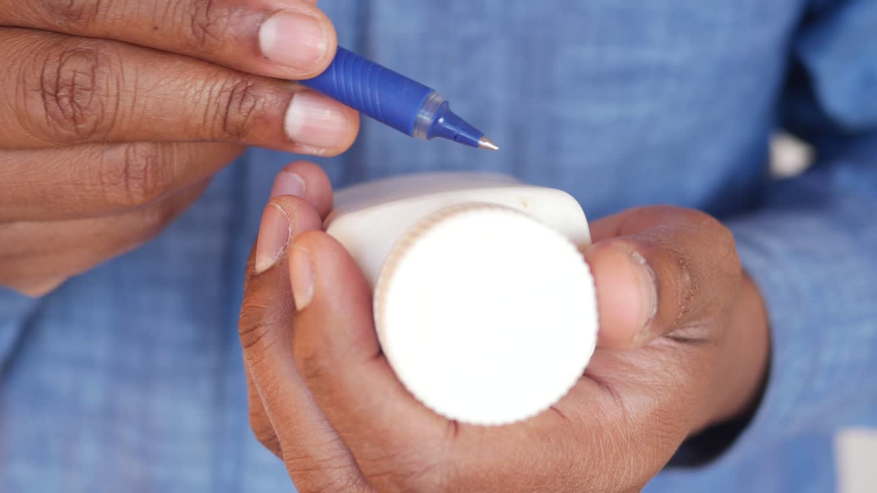 A person's hands holding a white bottle and a pen, appearing to write on it