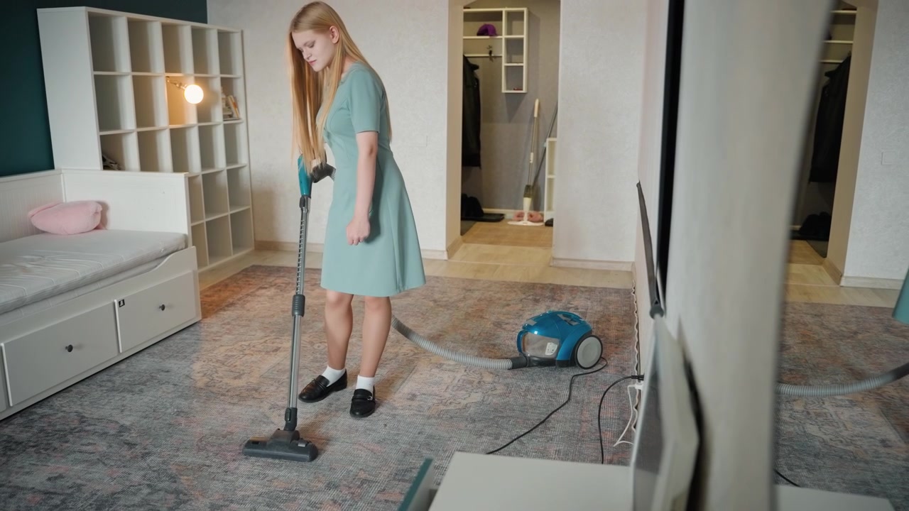 Ung vit kvinna dammsuger sovrum i hus. Metodisk dammsugning med cylinderdammsugare över mönstrad matta nära säng och hyllor. Lugn modern interiör, dagsljus, prydlig inredning, hushållssyssla, rutinunderhåll, fokuserad.