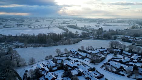 Aerial view of a quaint English village blanketed in fresh snow, with golden sunlight casting long shadows.