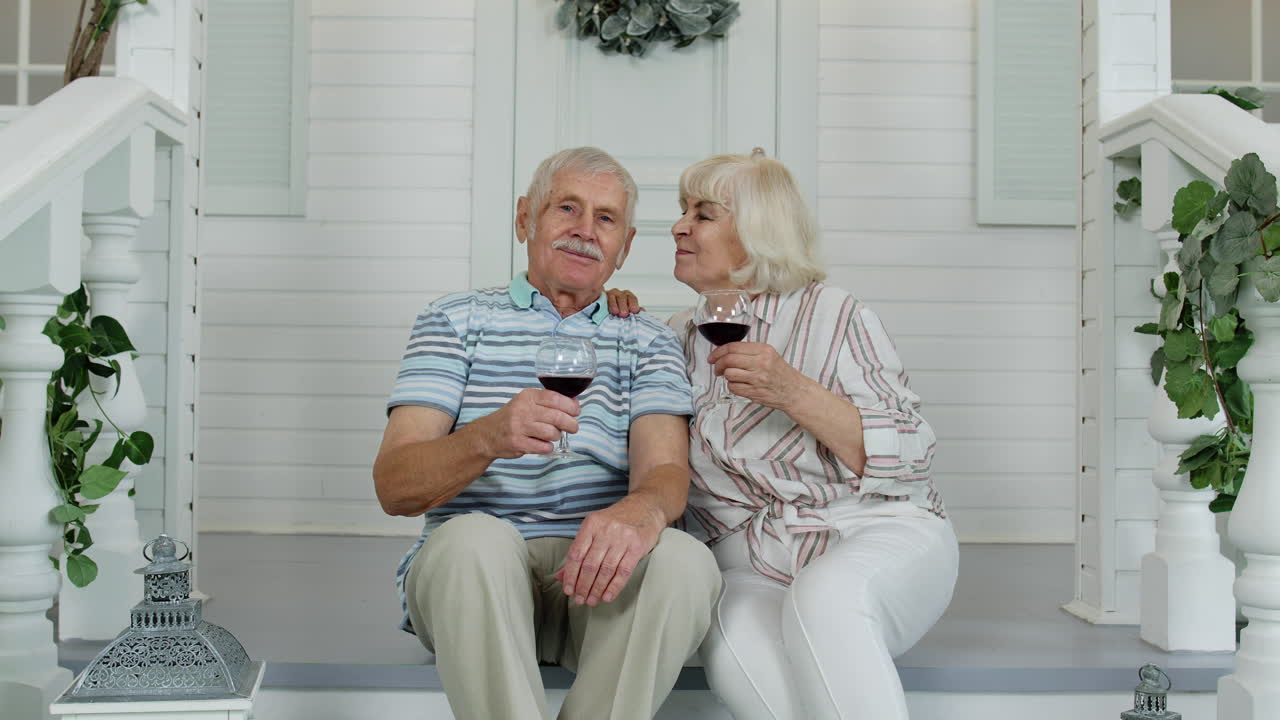 Attractive senior elderly Caucasian couple sitting and drinking wine in porch at home, making a kiss