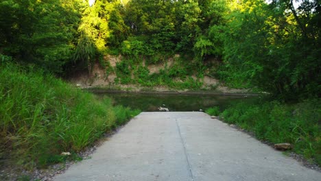 Drone-flying-or-really-steady-camera-walking-down-a-boat-ramp-towards-a-small-river-with-trees-on-all-sides