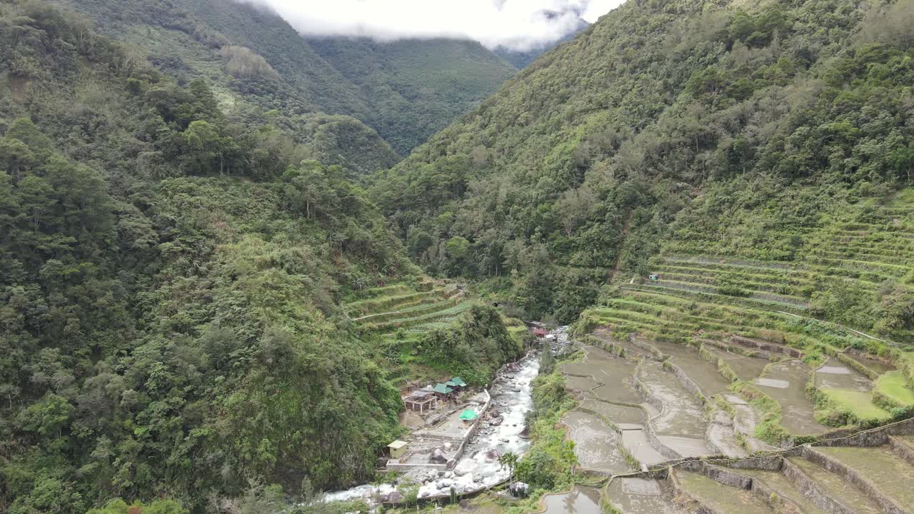 Aerial view of rice terraces on both sides of a rocky river in a lush valley in Northern Luzon, Philippines. Misty clouds hang over mountains in the background, creating a peaceful, natural landscape