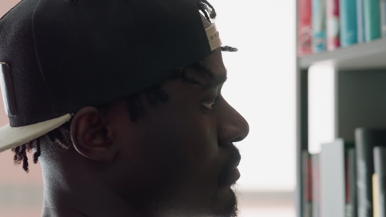 Close up side view of young man wearing cap with focused expression searching bookshelf in library, eyes scanning titles with intent concentration
