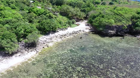 Aerial-backwards-flight-showing-private-beach-with-forest-palm-trees,-coral-reefs-and-rocks-underwater