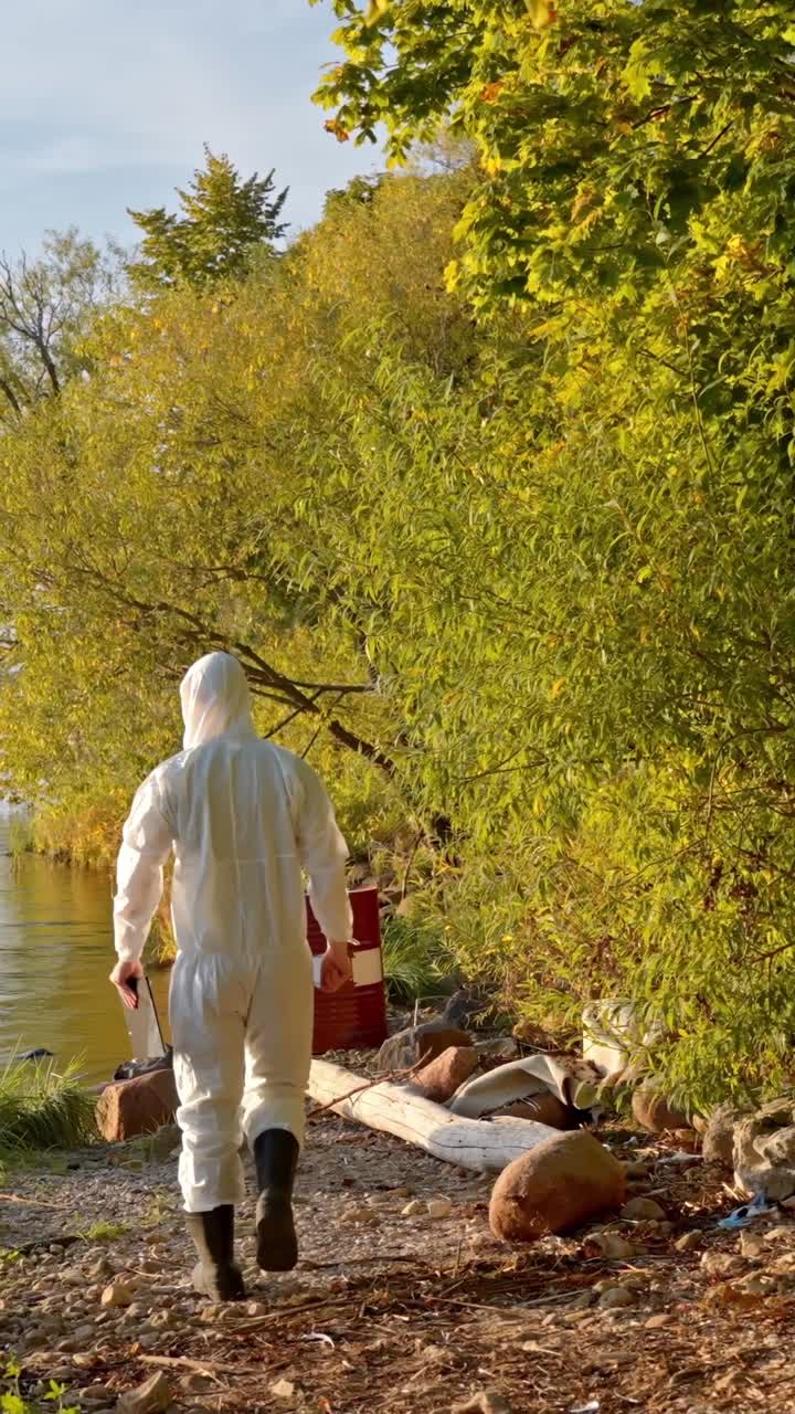 Scientist in white hazmat suit walks with clipboard near polluted lakeshore for inspection, vertical