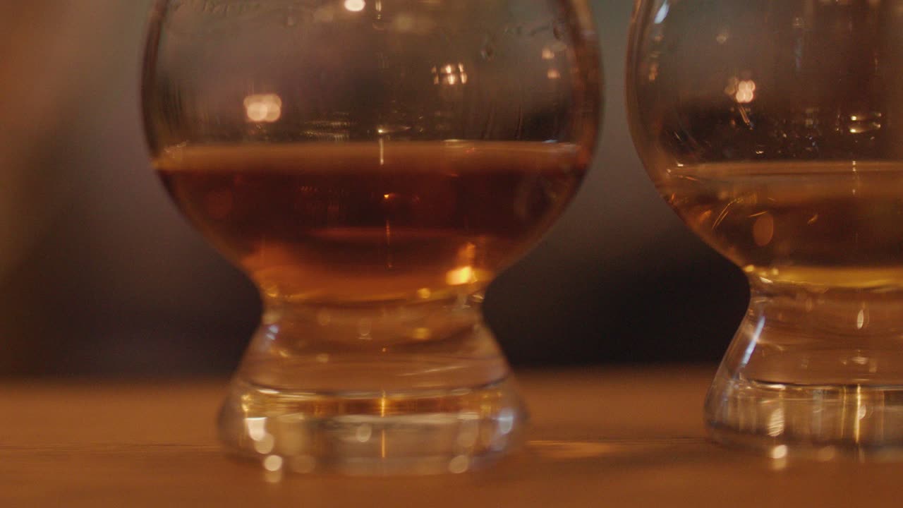 Two whisky glasses filled with amber liquid rest on a wooden table, softly illuminated by warm candlelight. Shallow depth of field and gentle camera movement create a cozy, intimate mood