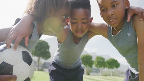 Video-of-three-african-american-schoolchildren-in-huddle-with-football-in-playing-field