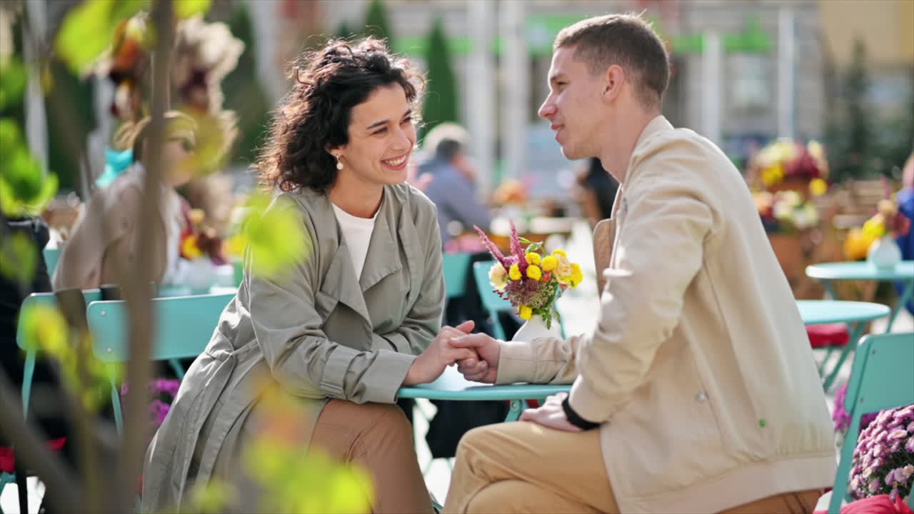 A romantic couple outdoors near a cafe. Holding hands, smiling, talking and looking at each other, flowers on the table. Autumn atmosphere. Slow motion
