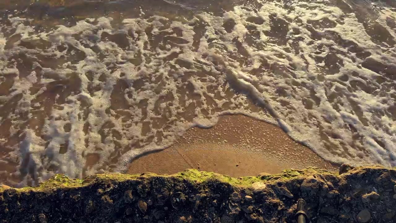 White ocean waves washing over smooth brown sand and algae covered rocks