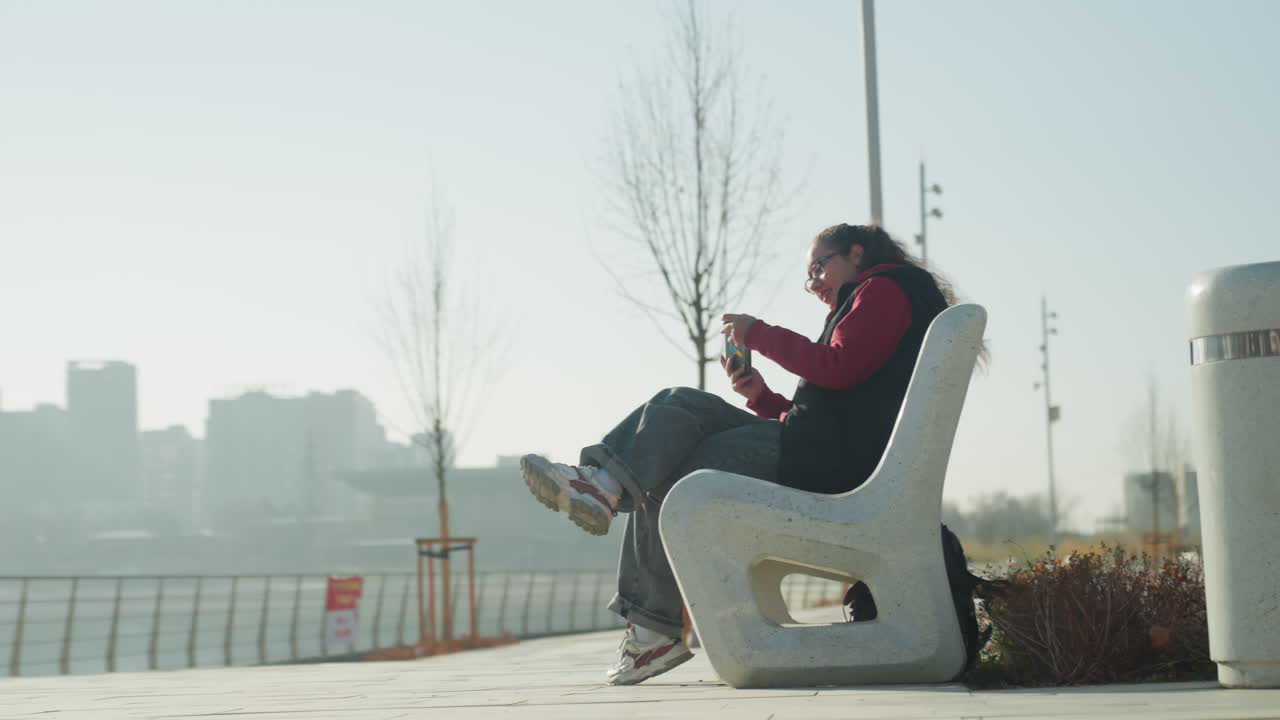Giovane donna su una panchina in riva al mare che scorre lo smartphone sotto la luce del sole, si stira giocosamente e ride dopo un messaggio, foschia sullo skyline, atmosfera rilassata da passeggiata, vibrazioni da viaggiatrice e studentessa.