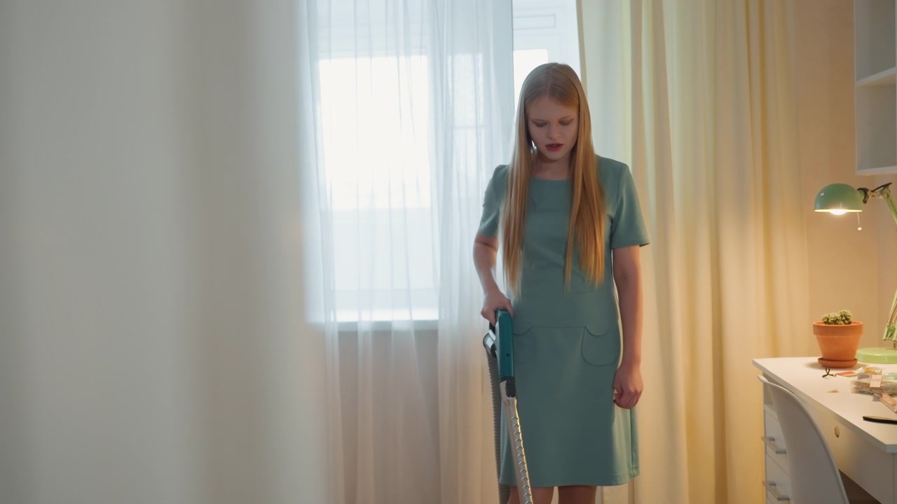 Donna caucasica che aspira la camera da letto vicino alla finestra, giovane proprietaria di casa in vestito verde si ferma a metà compito mentre pulisce la casa, luce mattutina soffusa attraverso tende trasparenti, scrivania con lampada e pianta in vaso visibili