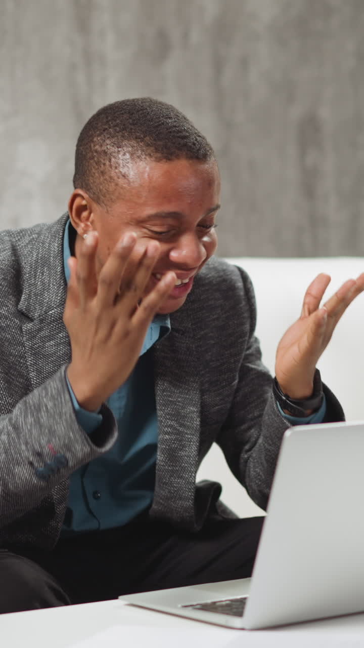 Positive African-American man tells funny story to colleagues at online meeting via modern laptop computer sitting at table in living room slow motion