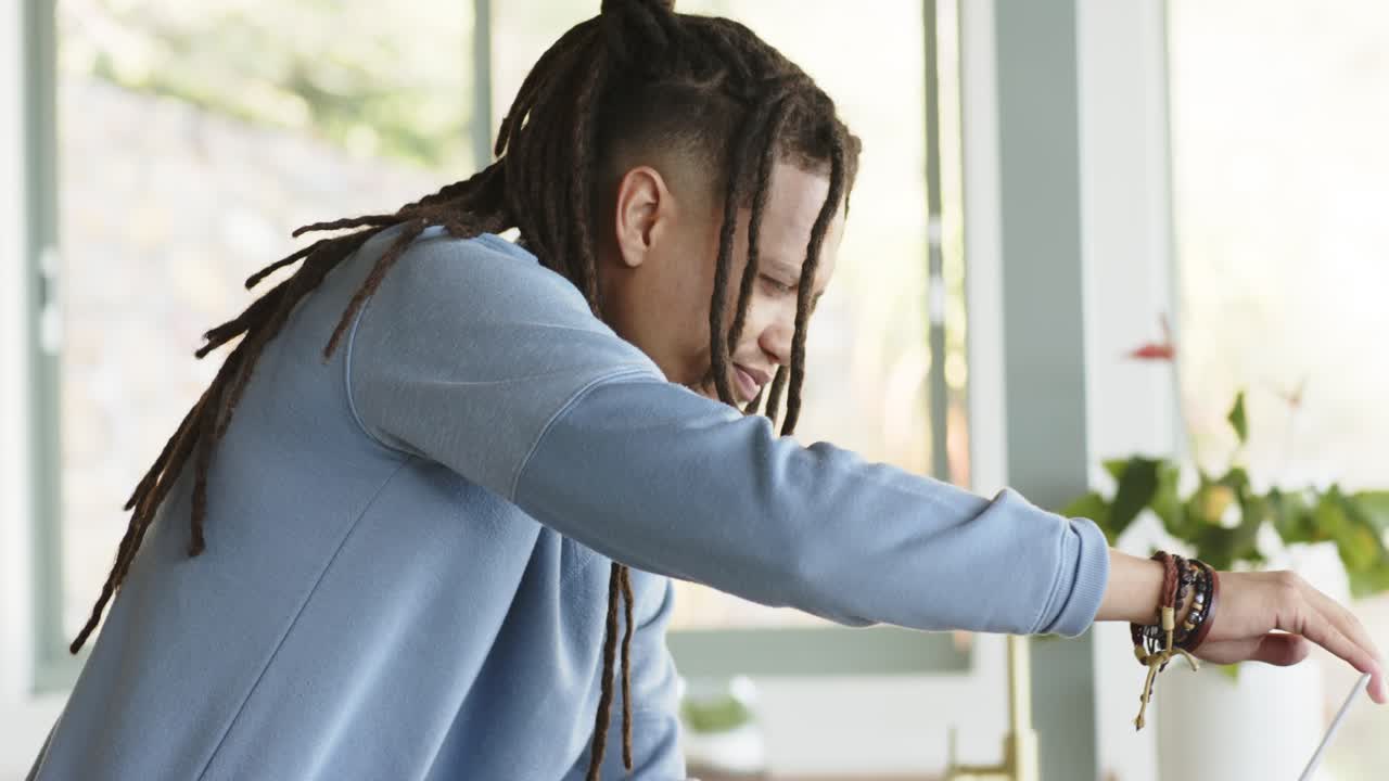 Man is leaning forward on countertop, initiating typing on silver laptop by window for work