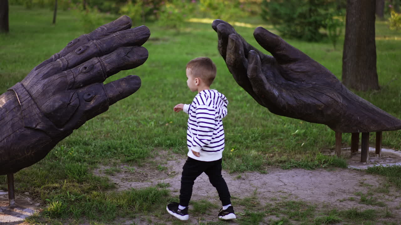 Little boy touches the wooden sculpture of big hands. Child walking in the green park in summer.
