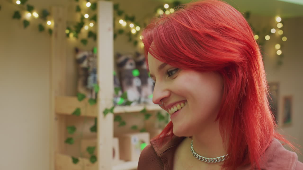 Mujer pelirroja de piel blanca sonriendo en una boutique rodeada de luces de hadas y plantas, sosteniendo una pasta envuelta, con un collar plateado, reacción sincera y alegre en sus roles de compradora y creadora, ambiente acogedor e íntimo