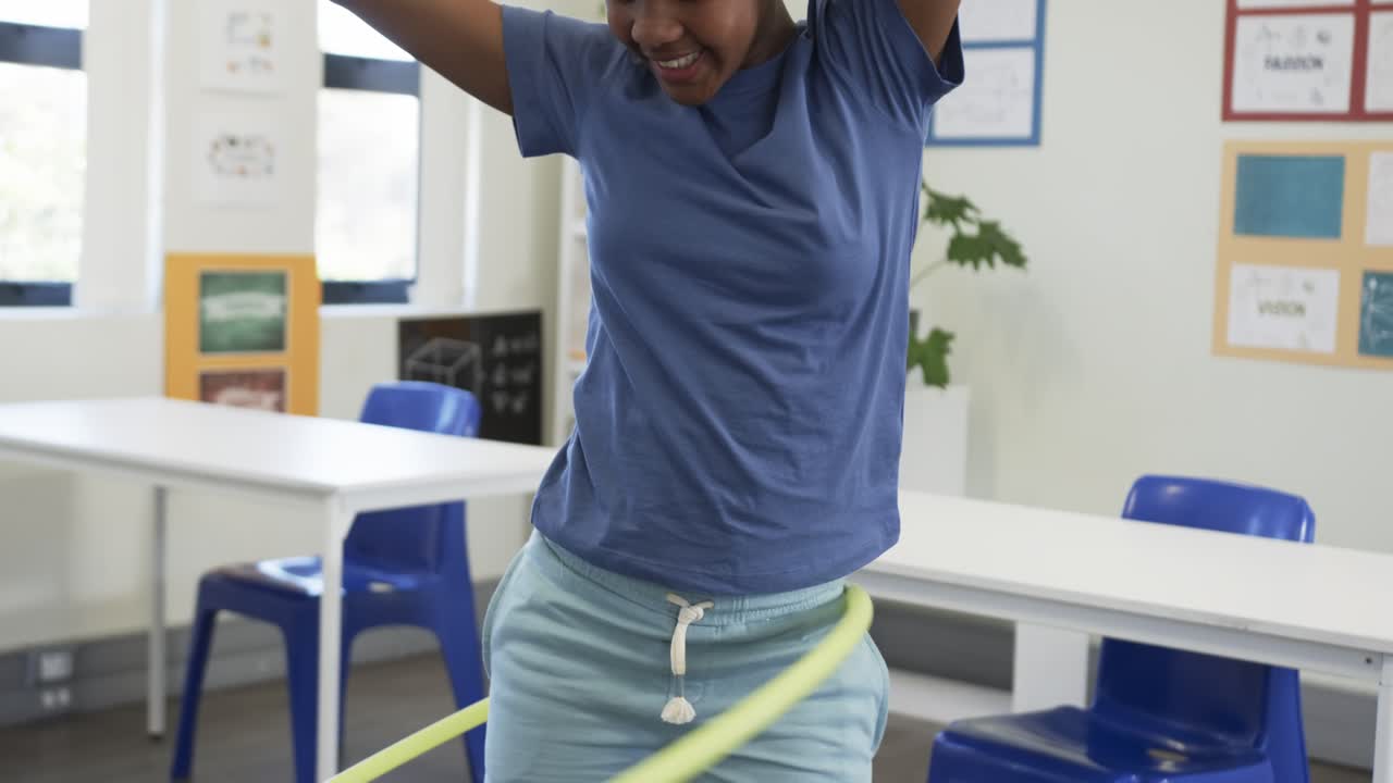 Child is entering frame and spinning yellow hula hoop at waist in classroom, practicing balance