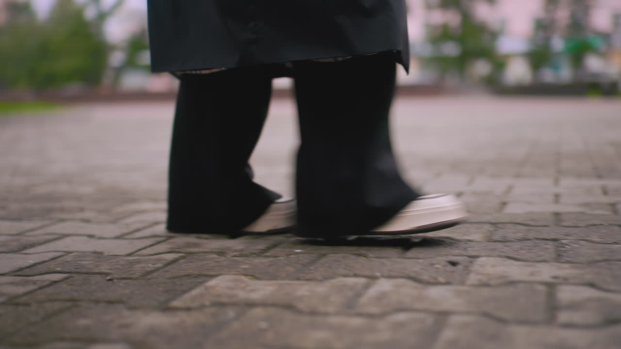 Lady walking on stone pavement wearing black pants and white sneakers, motion detail of steps outdoors during cloudy rainy day creating urban lifestyle atmosphere with blurred greenery in background