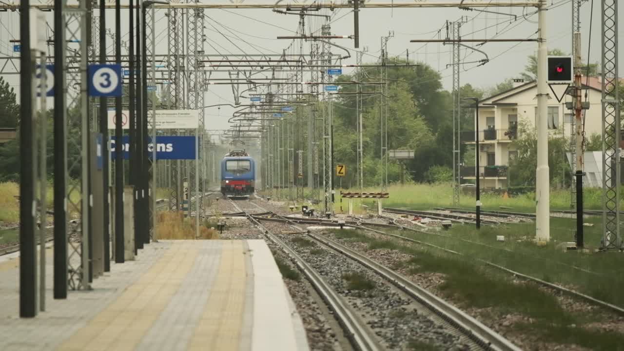 A train approaches a small countryside station surrounded by green fields under gentle falling rain