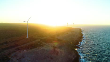 Golden-hour-glow-spread-across-wind-turbines-on-coastal-road-of-island,-drone-overview