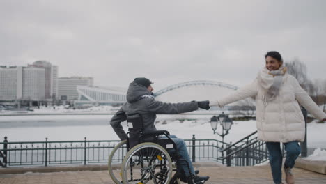 happy muslim woman and her disable friend in wheelchair spinning holding hands in city in winter