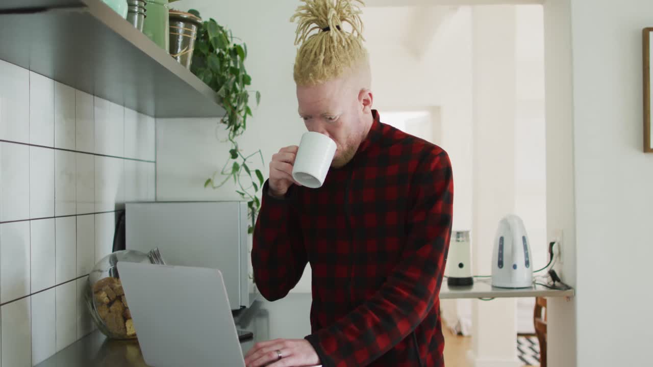 Homem afro-americano albino pensativo com dreadlocks usando laptop e tomando café