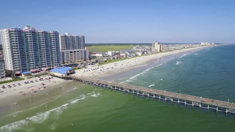 Drone-flying-over-ocean-towards-pier-and-beach