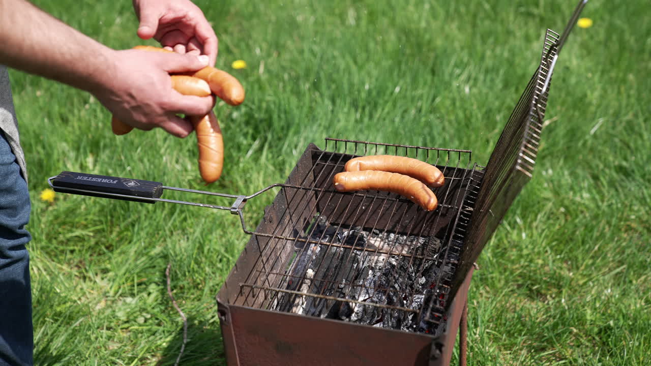 Uomo che mette le salsicce su una griglia per cuocerle. Cottura di salsicce hot dog sul fuoco. Grigliata all'aperto. Braciere su sfondo di erba verde