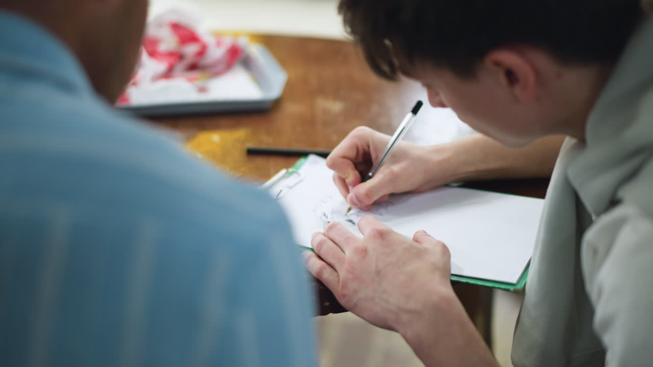 Youth wearing white polo shirt seated at wooden desk leaning over paper meticulously shading single spot to enhance contrast and beauty under soft natural light in creative workspace environment