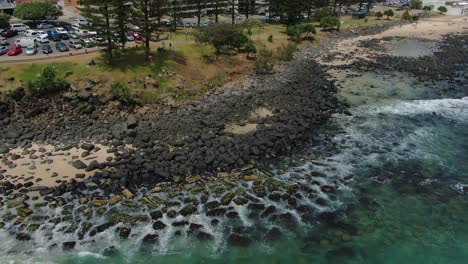 Descending-quickly-to-sea-level-over-rocky-beach,-bright-sunshine,-crashing-waves