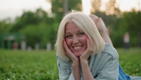 modern woman lying on green grass with hands under chin smiling gently, soft evening light, blurred trees behind, relaxed casual outdoor portrait capturing peaceful moment