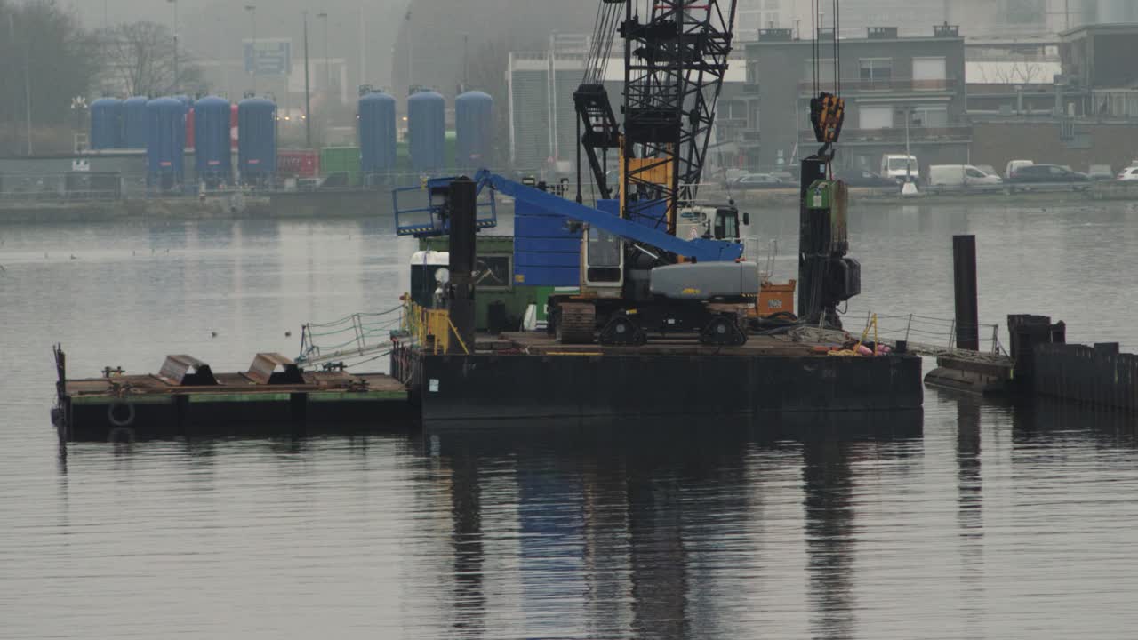 A crane and lift on a floating platform working on sheet pile installation in an industrial harbor