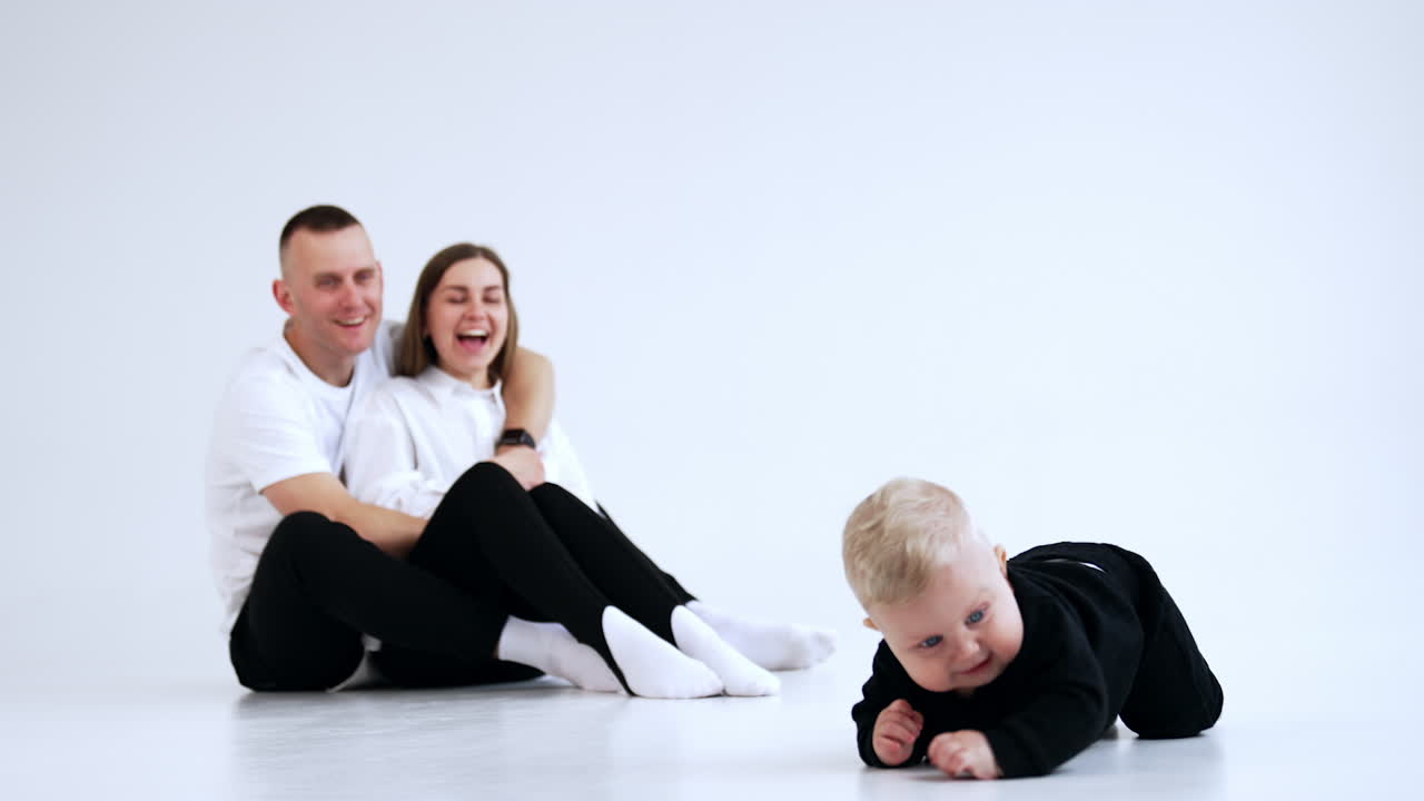 Little baby wearing black suit crawls by the floor. Smiling parents sit at backdrop watching their cute son. White backdrop.