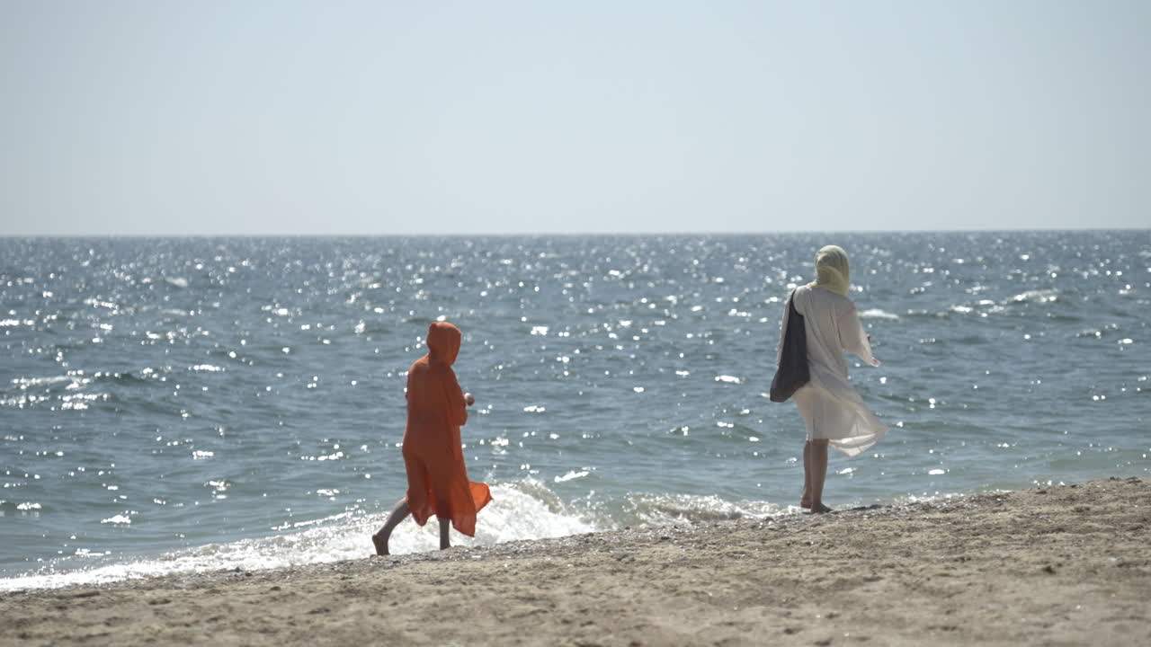 Rear view of two people walking on the beach in a summer sunny day.  Wind blowing in their light beach robes. Slow motion.
