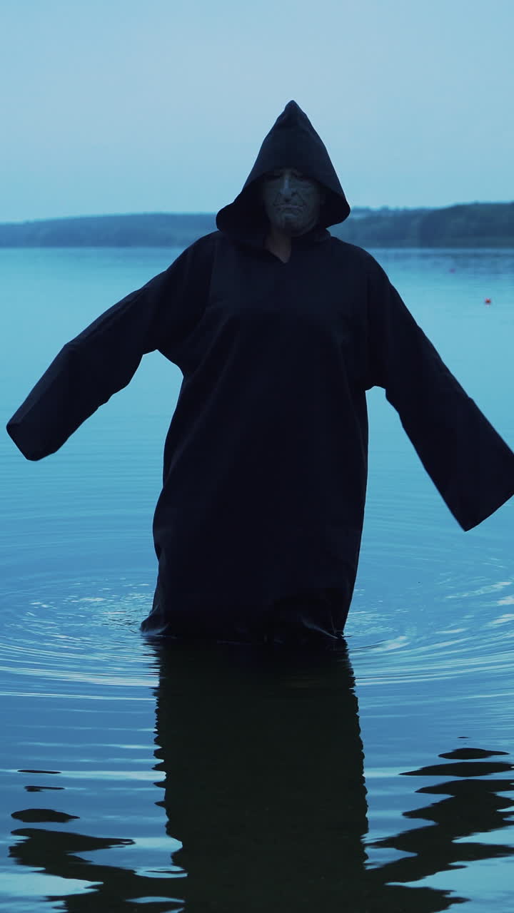Evil witch in black cloak with hood in the river. Halloween costume. Vertical video