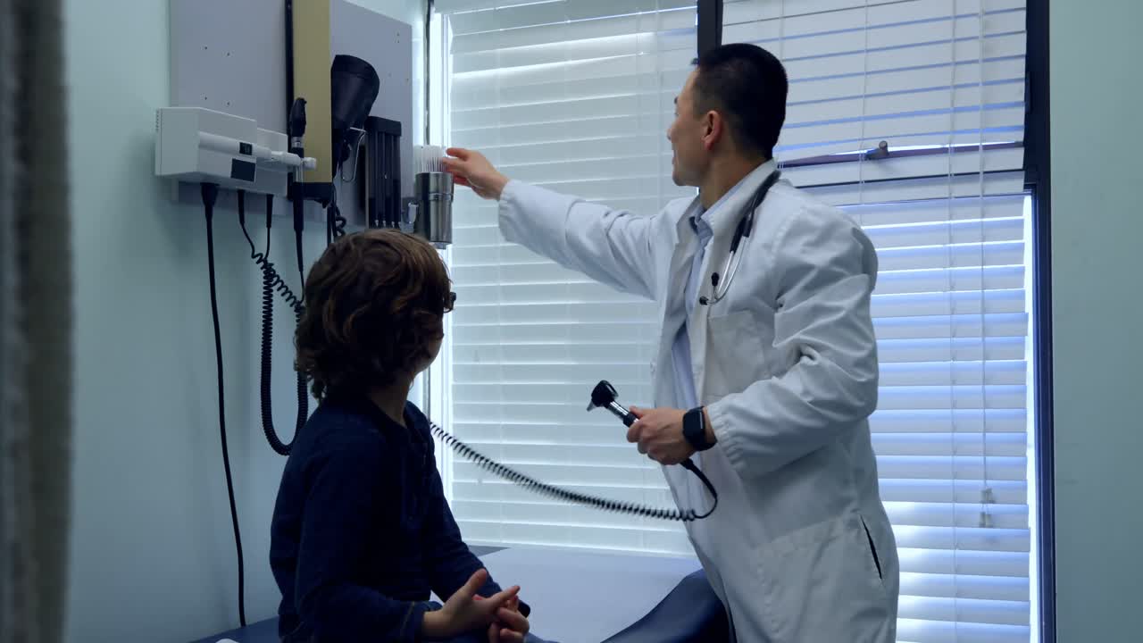Side view of young asian male doctor examining a caucasian boy patient ear with tool in a clinic 4k