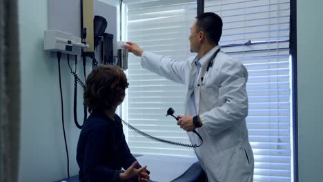 Side-view-of-young-asian-male-doctor-examining-a-caucasian-boy-patient-ear-with-tool-in-a-clinic-4k