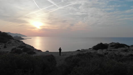 Awe-inspiring-drone-footage-captures-a-man-standing-on-edge-of-a-rocky-forest-cliff,-gazing-out-at-serene-sea-as-golden-hour-sunrise-breaks-before-his-eyes