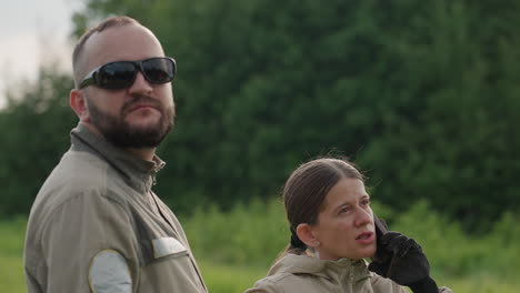 colleague duo stand in lush green field observing sky signals while woman makes urgent phone call reporting aerial device status to team using handheld controller and smartphone