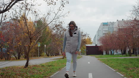 confident woman walking in sport outfit during summer on urban road with wind gently blowing autumn leaves as trees sway around modern buildings with cars in background