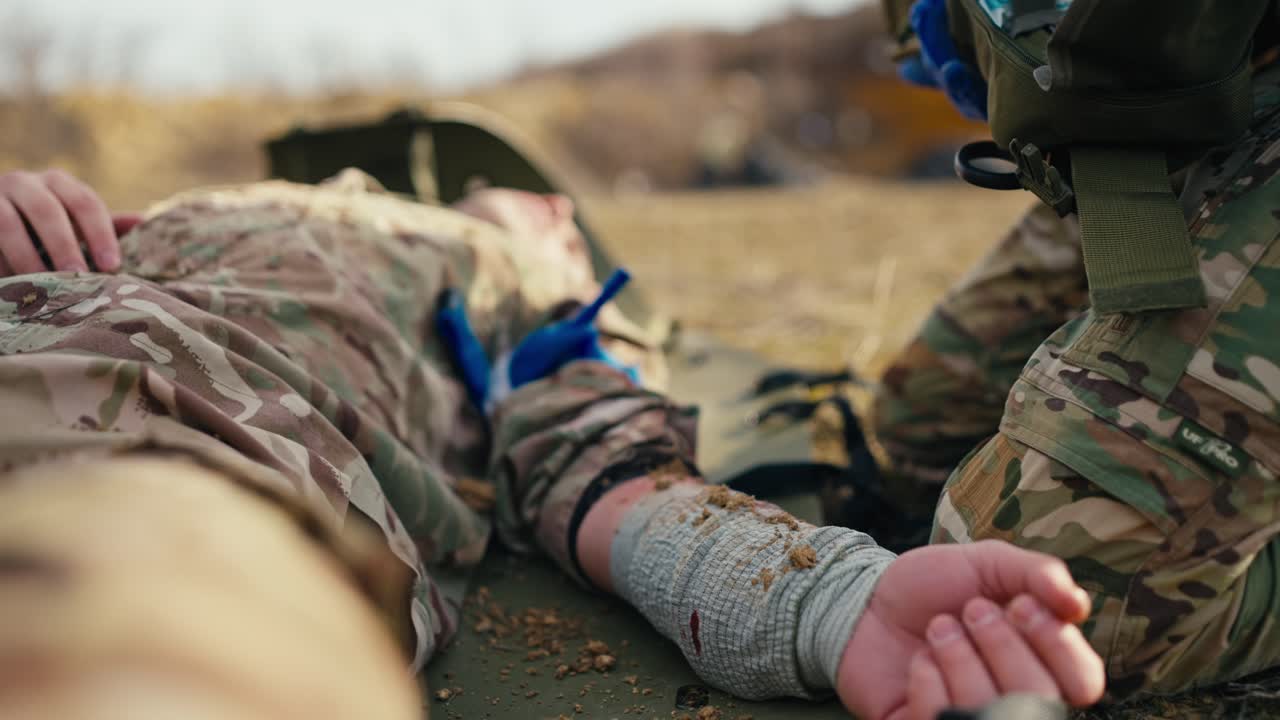 Close-up a military man with his arm bandaged with a cloth bandage lies unconscious on an army stretcher while a combat medic near him looks for a special medicine in his first aid kit during combat operations at the training ground