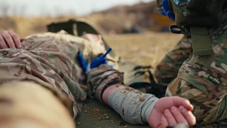 close-up a military man with his arm bandaged with a cloth bandage lies unconscious on an army stretcher while a combat medic near him looks for a special medicine in his first aid kit during combat operations at the training ground