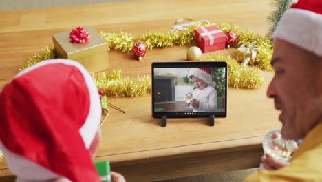 Padre-E-Hijo-Caucásicos-Con-Gorros-De-Papá-Noel-Usando-Una-Tableta-Para-Una-Videollamada-Navideña-Con-Un-Niño-En-La-Pantalla