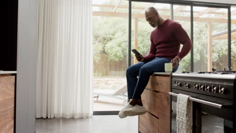 Busy-mature-african-american-man-sitting-on-countertop-using-smartphone-in-kitchen,-slow-motion