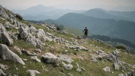 Excursionista-Caminando-Hacia-La-Cámara-En-Una-Colina-Con-Bastones-De-Senderismo,-Valle-Y-Otras-Montañas-Visibles-En-El-Fondo,-Parcialmente-Nublado