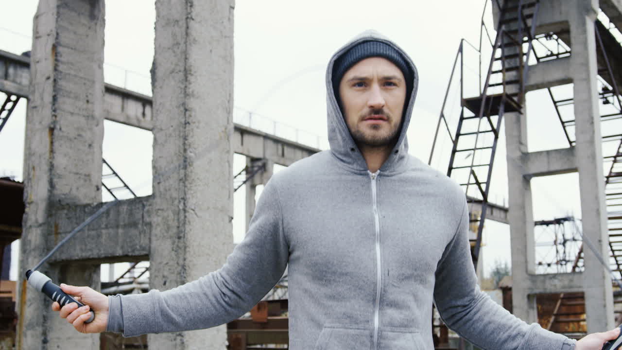 retrato de un deportista caucásico con barba en una capucha gris saltando la cuerda al aire libre en una fábrica abandonada en una mañana nublada