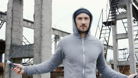 portrait of caucasian sportsman with beard in a grey hoodie jumping rope outdoors an abandoned factory on a cloudy morning