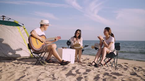 group of happy asian friends playing guitar and singing with clap while picnic and camping on the beach in outdoors vacation summer.