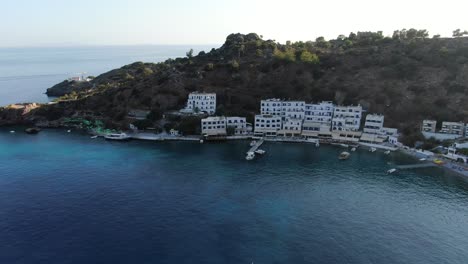 Drone-view-in-Greece-flying-over-blue-sea-in-Loutro-small-white-house-town-and-small-boats-next-to-a-hill-on-a-sunny-day